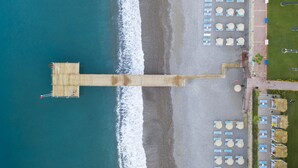 Vista aérea de un hotel frente a la playa con un muelle que se adentra en el mar, hileras de sillas de playa y sombrillas.