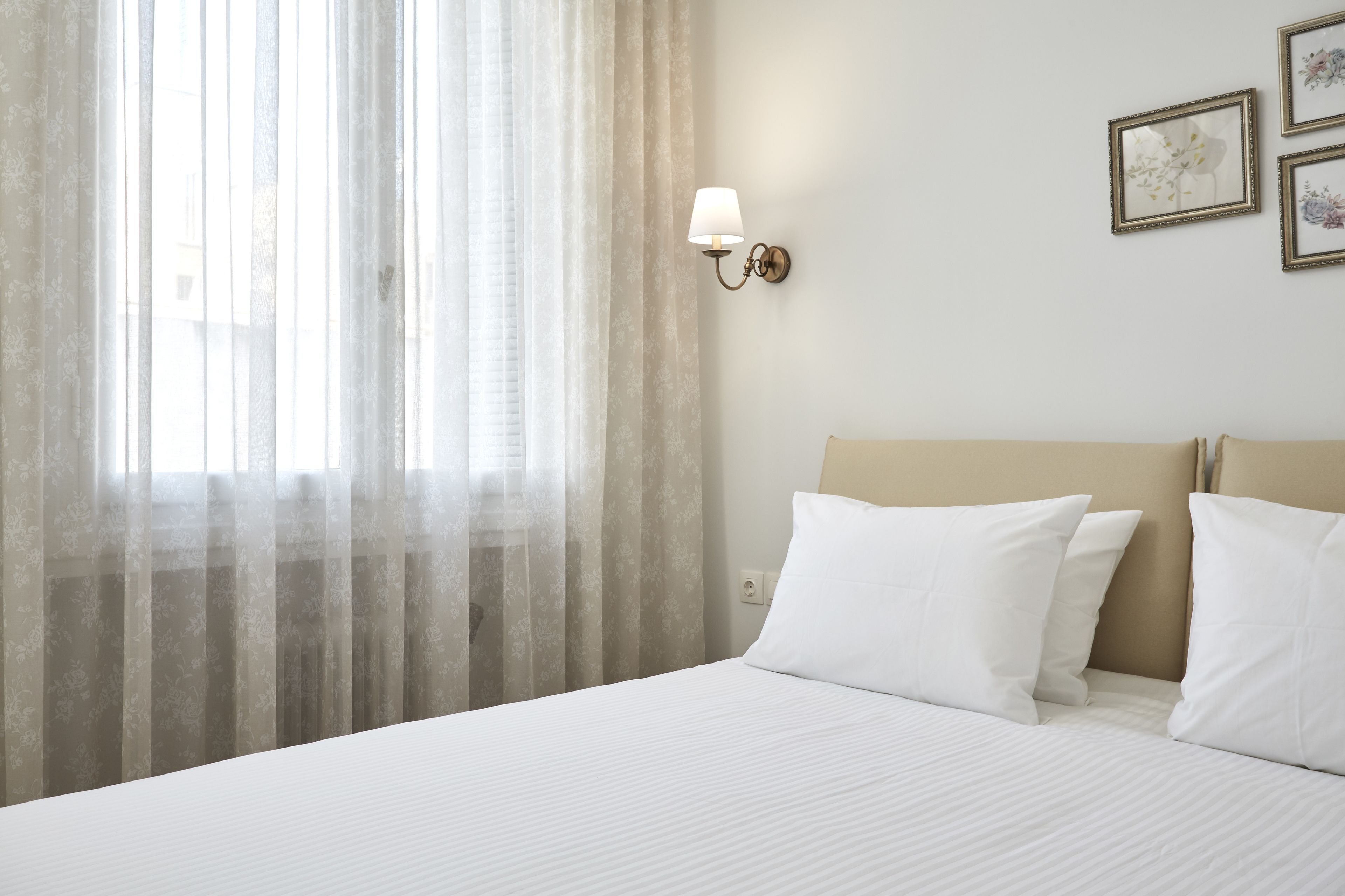 Een hotelkamer met een bed, dunne gordijnen, een wandlamp en kunstwerken in lijsten.