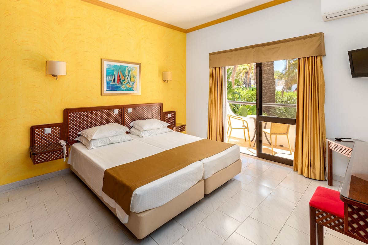 A hotel room with a large bed, a painting on the wall, and a balcony with yellow curtains.
