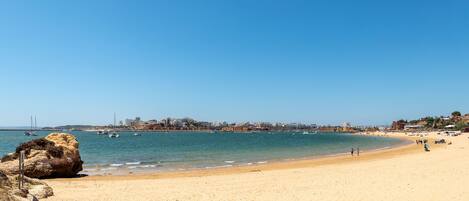 A wide sandy beach with a clear blue sky, calm sea, and a distant cityscape with buildings and boats.