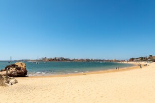 Una extensa playa de arena clara, cielo azul despejado, mar tranquilo y un horizonte lejano con edificios y barcos.
