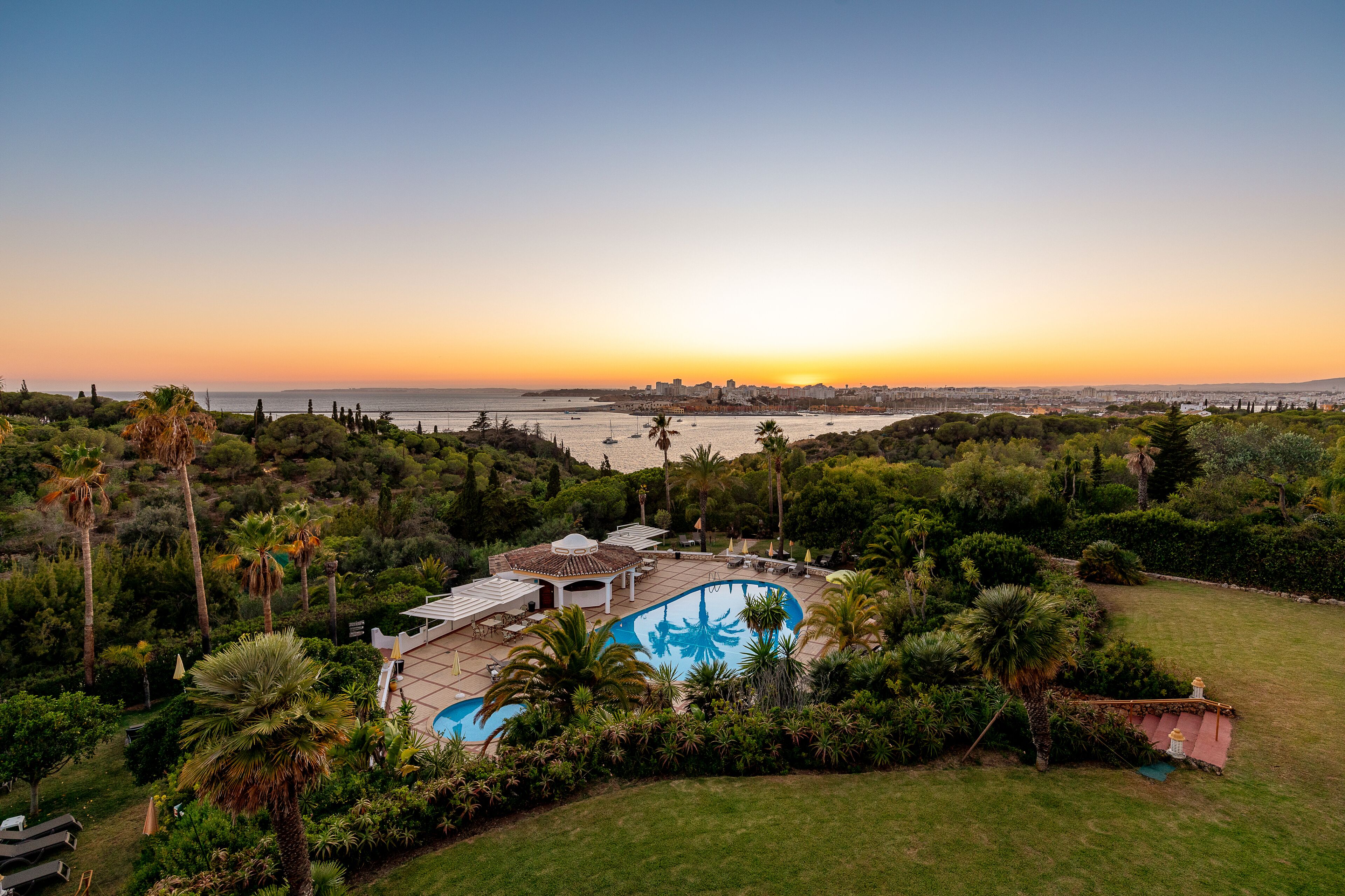 Una gran piscina envoltada de vegetació exuberant, amb vista a un paisatge urbà i l'oceà al capvespre.