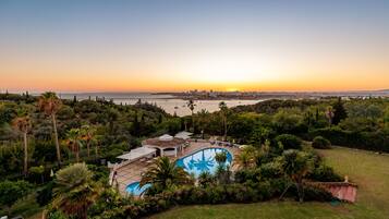 A large swimming pool surrounded by lush greenery, with a view of a cityscape and the ocean at sunset.