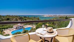 A balcony with wicker chairs, a small table with a book and a glass of drink, and a view of a swimming pool and cityscape. - Hotel Casabela (Lagoa)