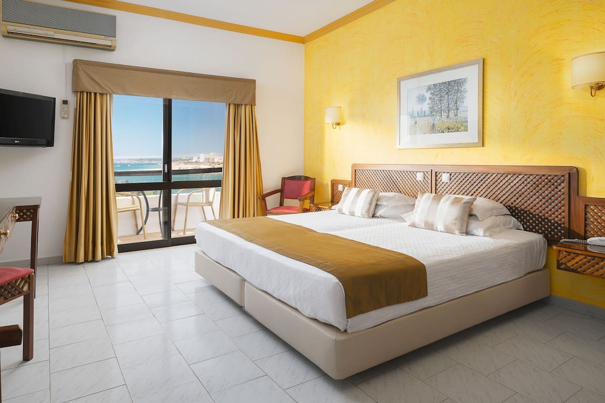 A hotel room with a large bed, a TV, a desk with a lamp, a chair, and a balcony with a view of the sea.