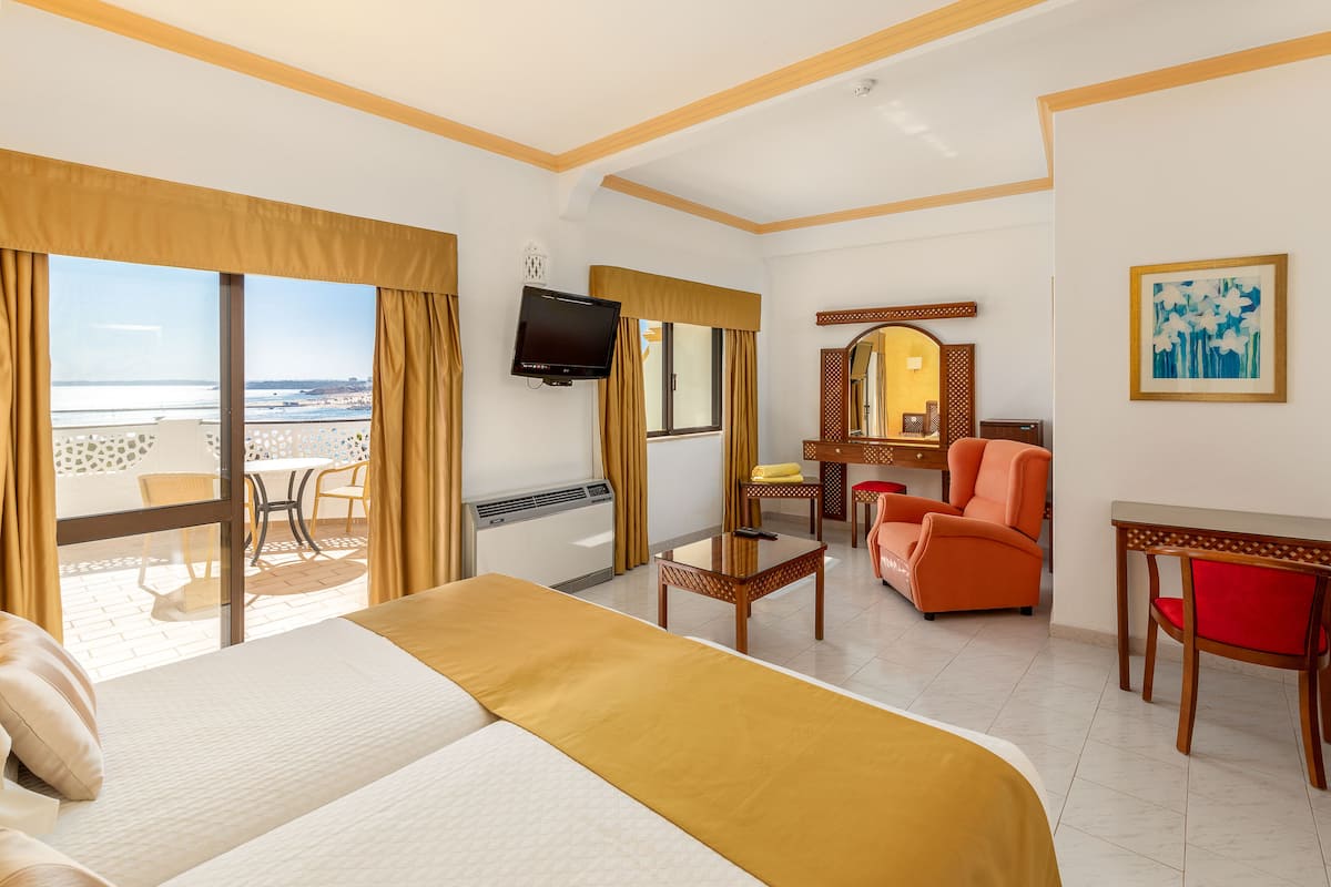 A hotel room with a bed, a desk, a chair, a TV, and a view of the beach.
