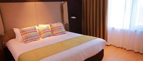 A neatly made bed with a white comforter and a yellow throw blanket, two colorful geometric pillows, and a headboard.