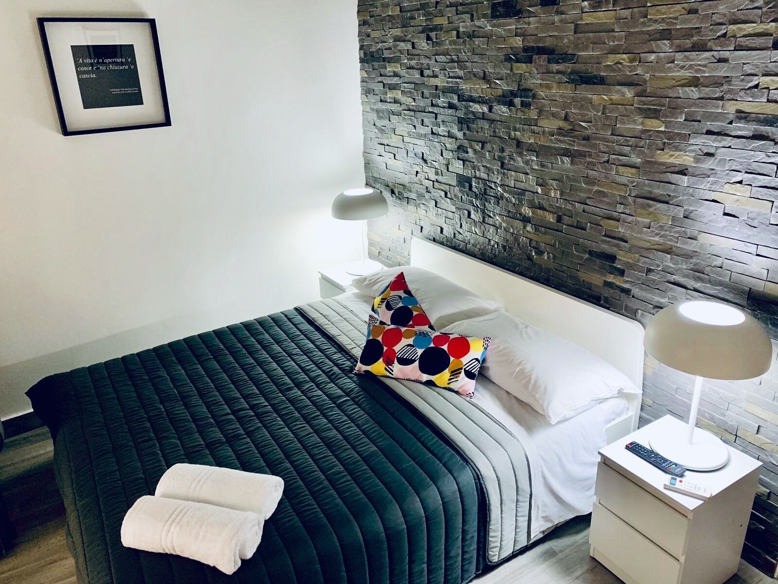 A neatly arranged hotel room with a bed, a nightstand, a lamp, and a wall with a stone pattern.