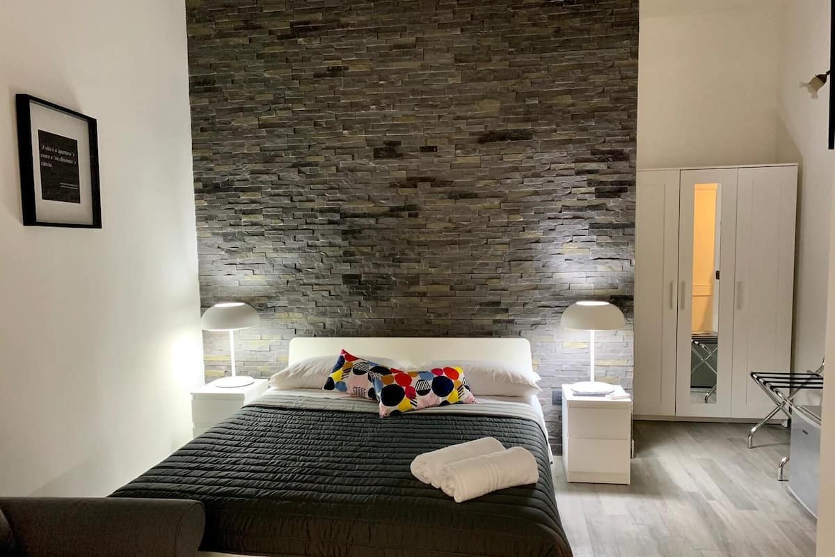 A modern bedroom with a stone wall, a bed with a colorful pillow, two bedside lamps, and a wardrobe with a mirror.