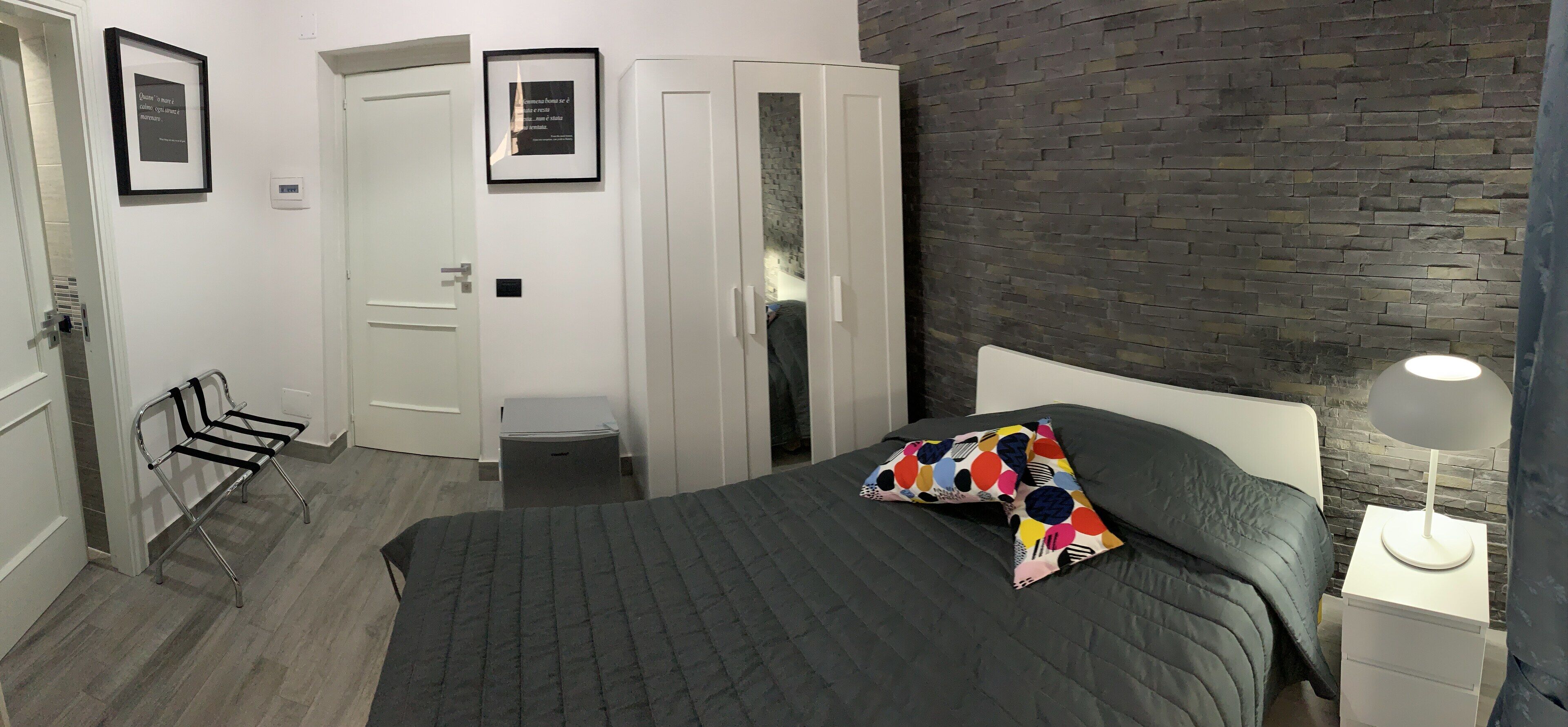 A bedroom with a bed, a nightstand, a chair, a wardrobe, and a wall with a brick pattern.