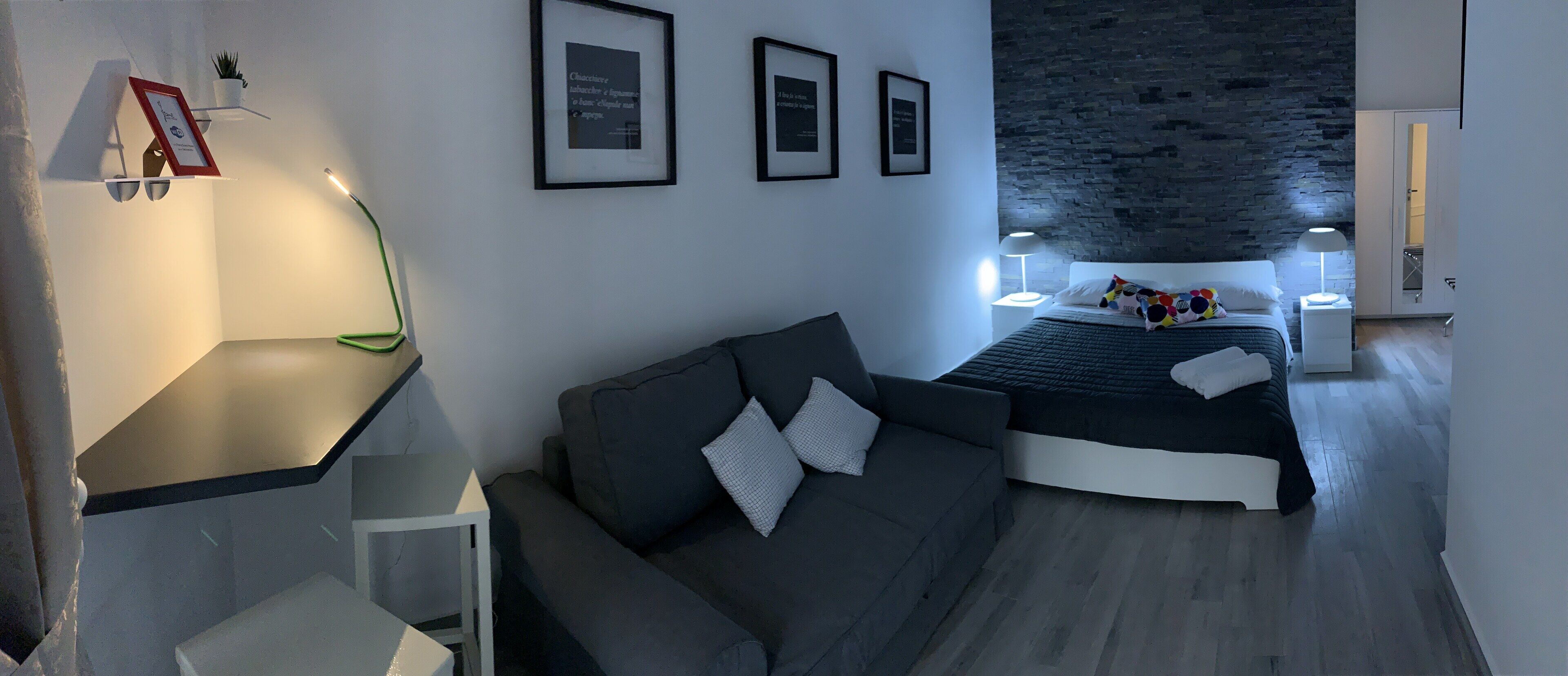 A modern bedroom with a bed, a gray sofa, and a small table. There are framed pictures on the wall and a plant on a shelf.