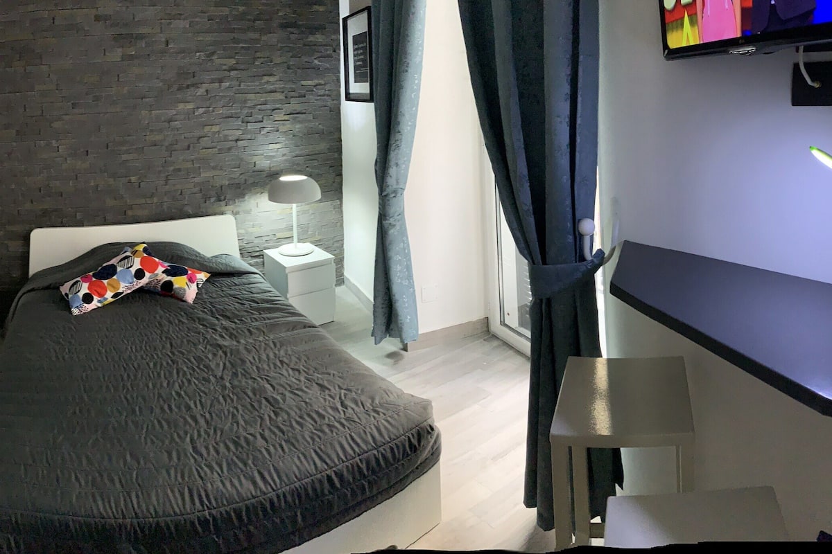 A compact bedroom with a bed, a small bedside table with a lamp, a white wardrobe, and a grey refrigerator.