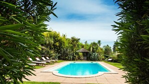 Una piscina rectangular con borde de baldosas, rodeada de exuberante vegetación y un pabellón.
