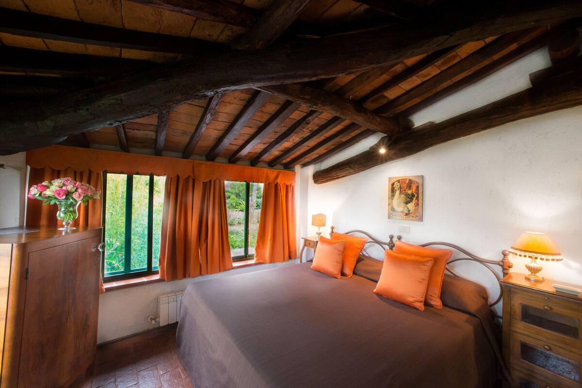 A bedroom with a wooden ceiling, a large bed with orange bedding, a nightstand with a lamp, and a window with curtains.