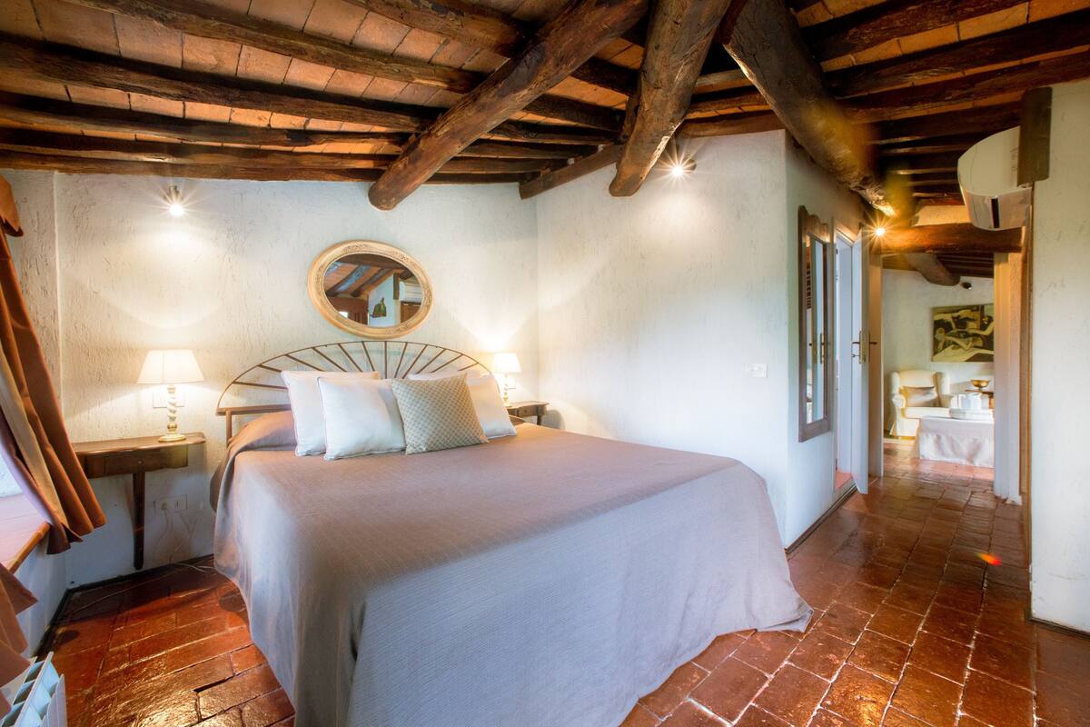 A bedroom with a large bed, a wooden ceiling, a round mirror, and a tiled floor.