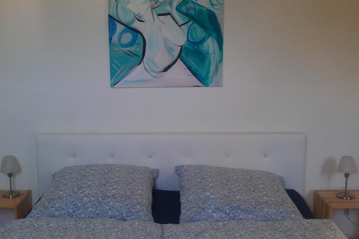 A bedroom with a white headboard, two patterned pillows, a painting on the wall, and two bedside lamps.