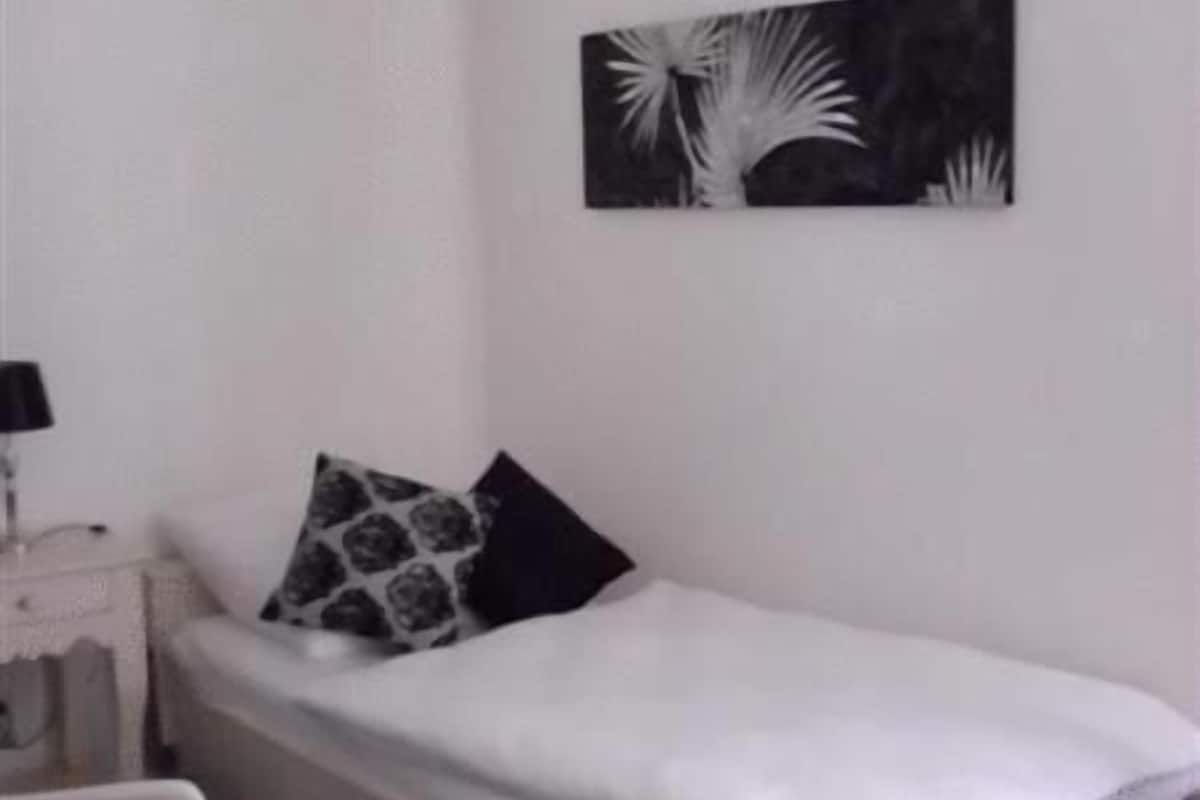 A single bed with a white mattress and a black and white pillow, a small bedside table with a lamp, and a wall-mounted artwork.
