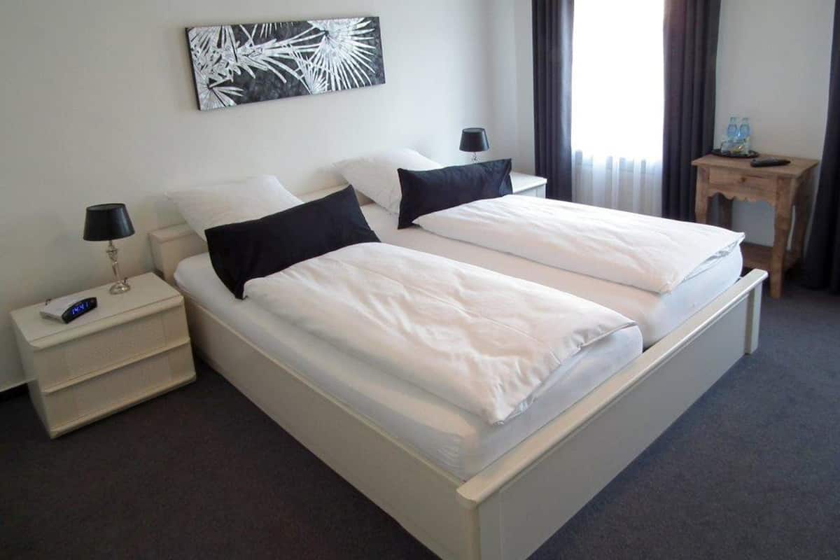 A double bed with white bedding, a bedside table with a lamp, a small wooden side table with water bottles, and a wall-mounted abstract art piece.