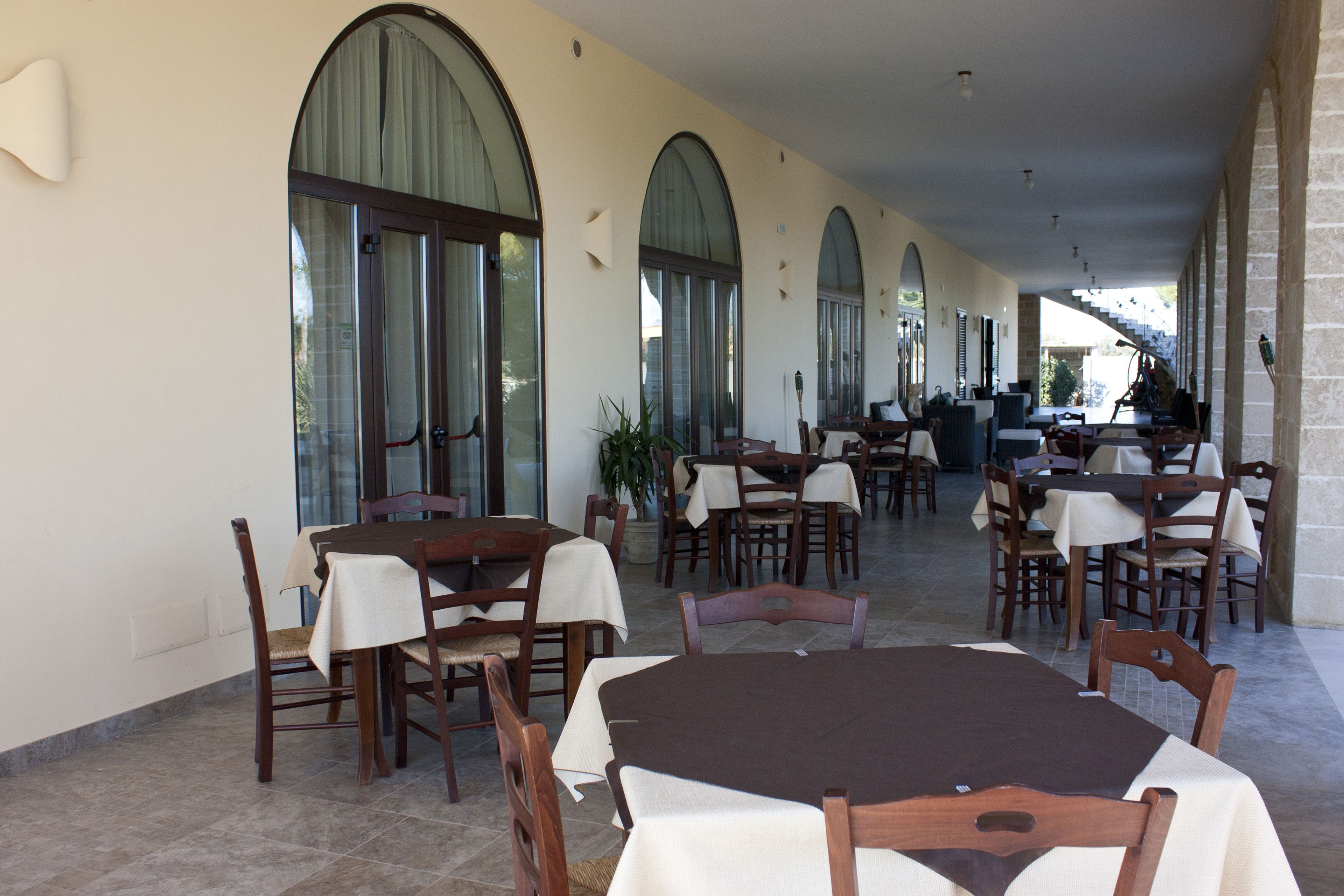 A covered outdoor dining area with multiple tables and chairs, large arched windows, and a stone wall on one side.