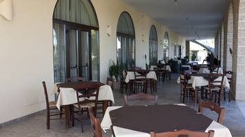 A covered outdoor dining area with multiple tables and chairs, large arched windows, and a stone wall on one side.