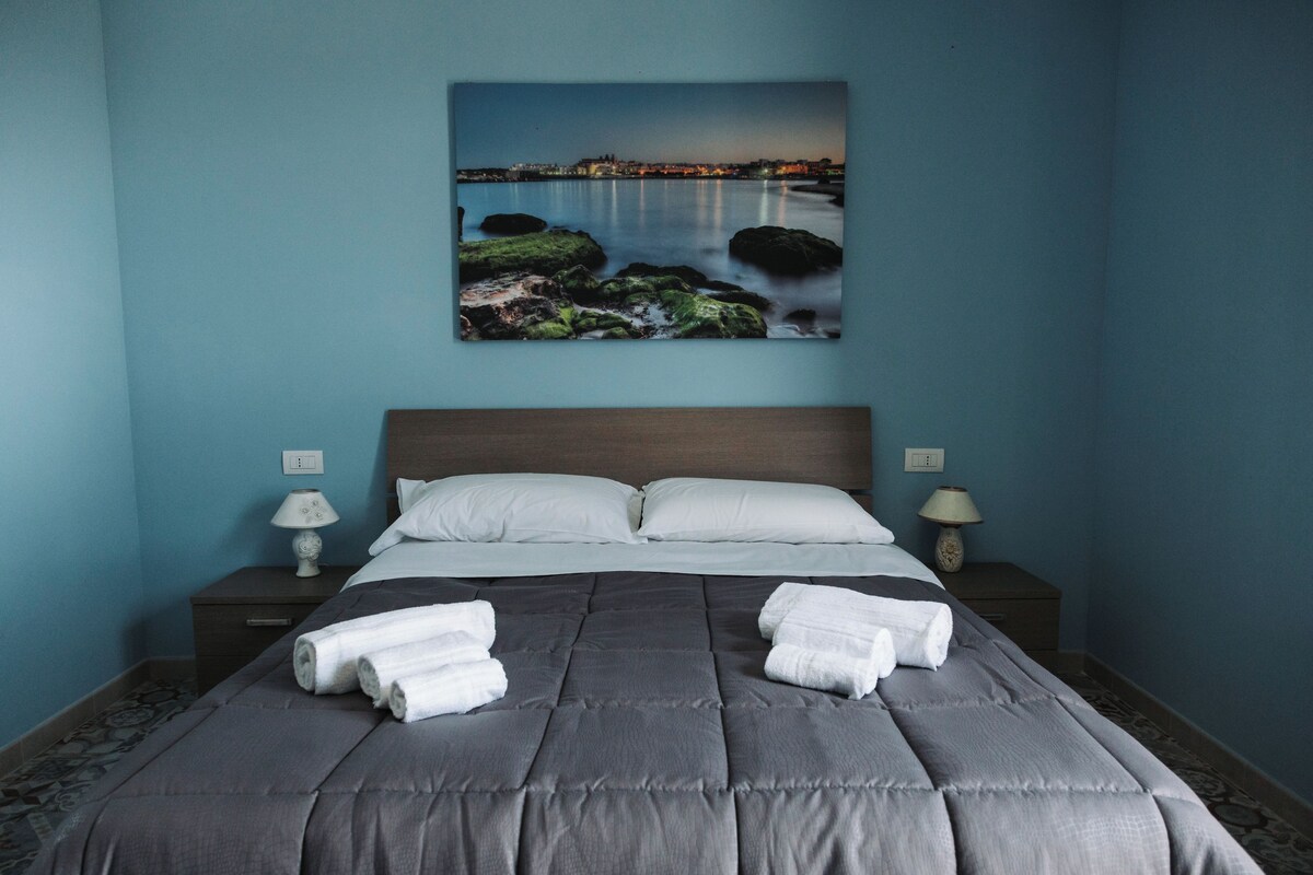 Un dormitorio con cama, dos mesitas de noche, un cuadro de una escena costera y toallas sobre la cama.