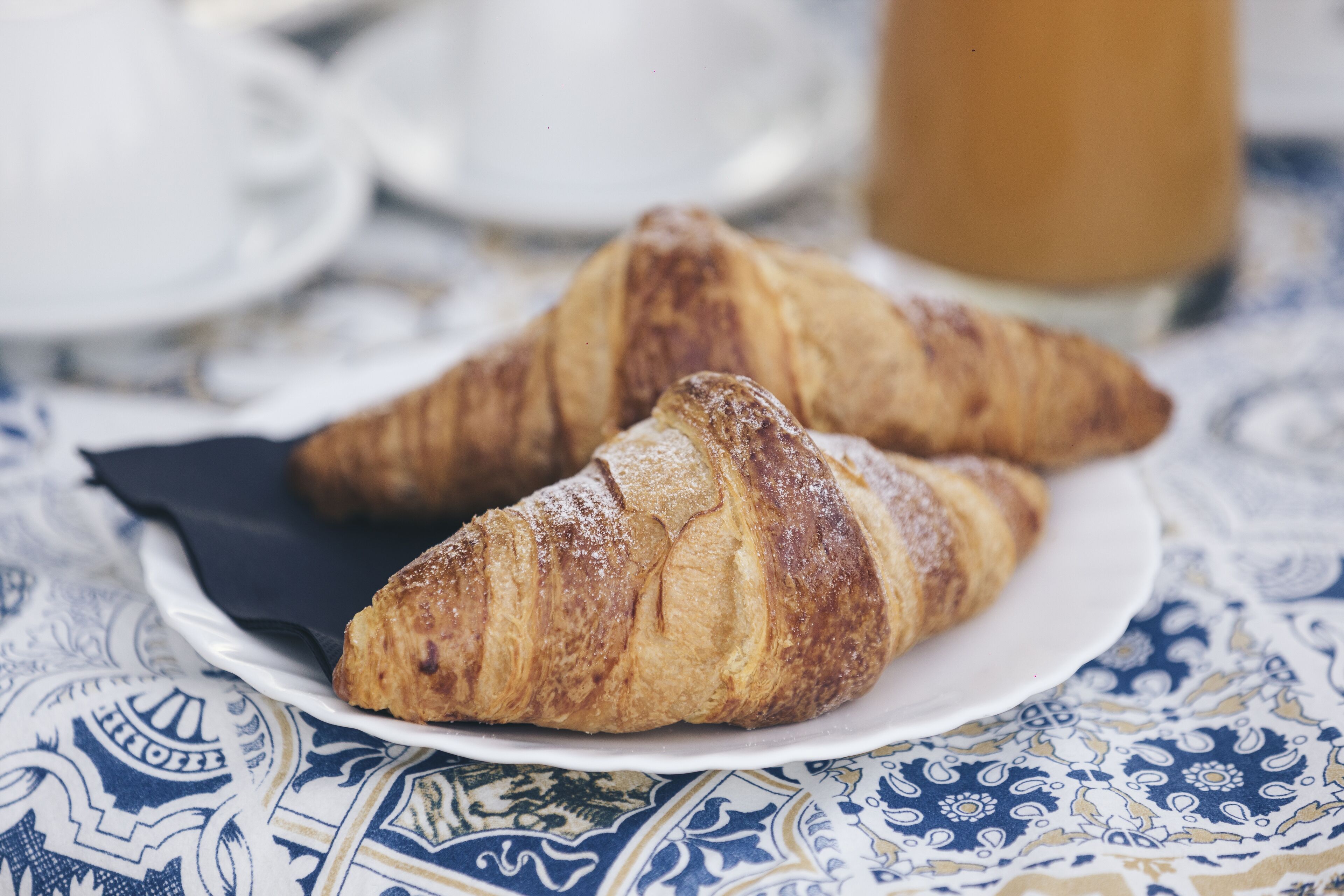 Zwei Croissants auf einem weißen Teller mit gemustertem Tischtuch.