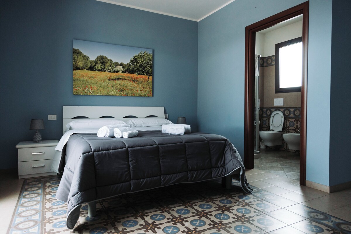 A bedroom with a bed, a nightstand, a wall-mounted painting, and a bathroom visible through an open door.