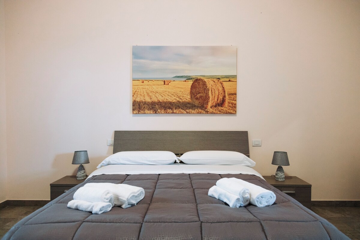 Habitación de hotel con cama, dos mesitas de noche con lámparas, un cuadro de un campo con pacas de heno y toallas enrolladas sobre la cama.