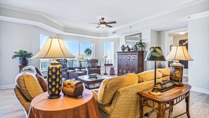 A spacious living room with a large sofa, a coffee table, and two lamps. There is a ceiling fan and a view of the outdoors through large windows.