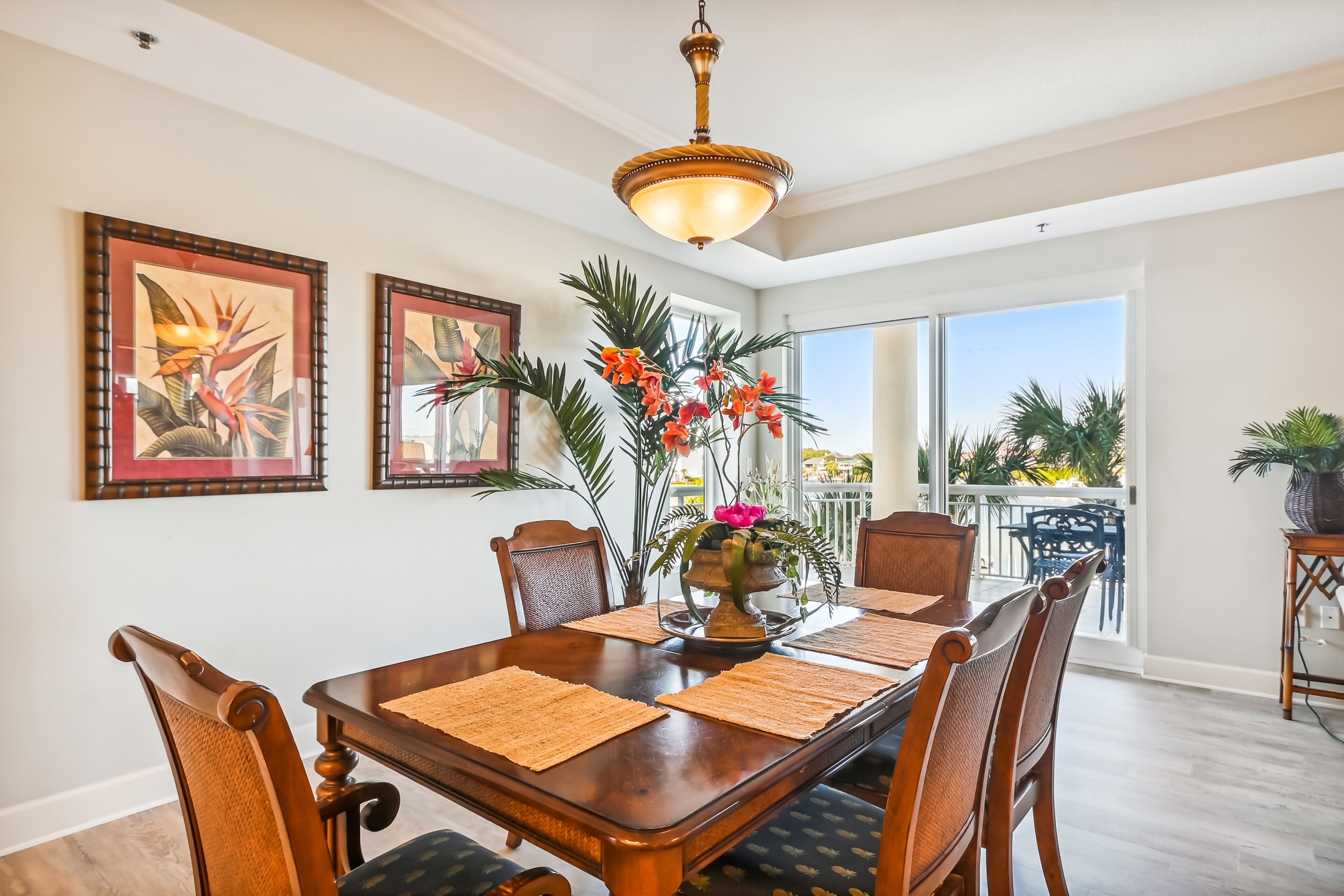 A dining area with a wooden table, chairs, and a vase with flowers. There are two framed pictures on the wall and a ceiling light fixture above the table.