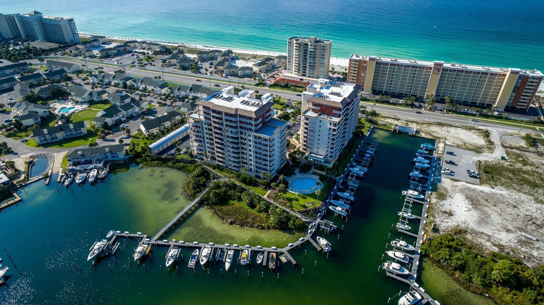 Vista aérea de uma marina costeira com vários barcos atracados, rodeada por edifícios residenciais e um hotel.