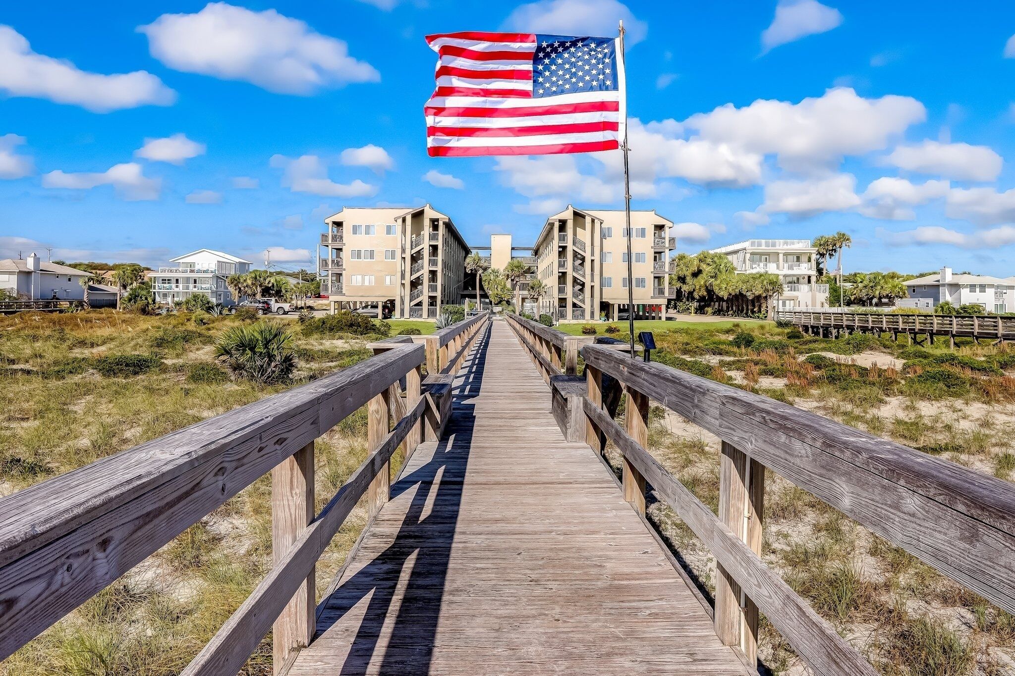 A wooden boardwalk leading to a building complex with multiple balconies and a flagpole with an American flag.
