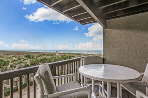 A balcony with a white table and chairs, overlooking a coastal landscape with palm trees and a view of the ocean. - Ocean View Condo Steps Away from Beach, Access to Salt Water Pool by RedAwning (Fernandina Beach)