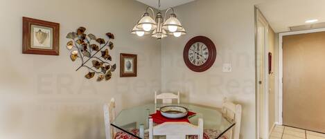 Un comedor con mesa de vidrio, sillas blancas, una lámpara de araña y adornos en la pared, incluyendo un reloj y cuadros enmarcados.