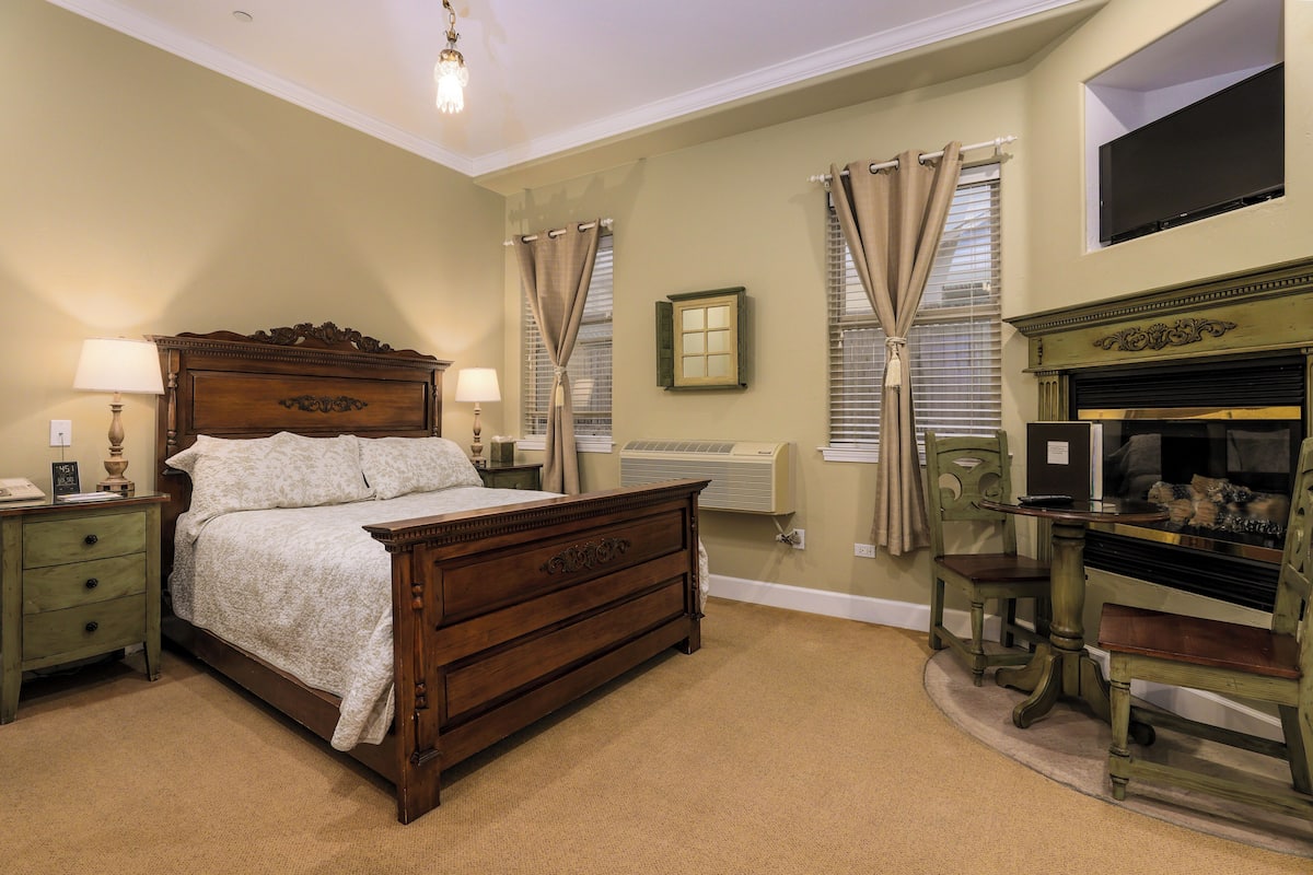 Une chambre à coucher avec un lit en bois, une table de chevet, une commode, une chaise, une cheminée et une fenêtre avec des rideaux.