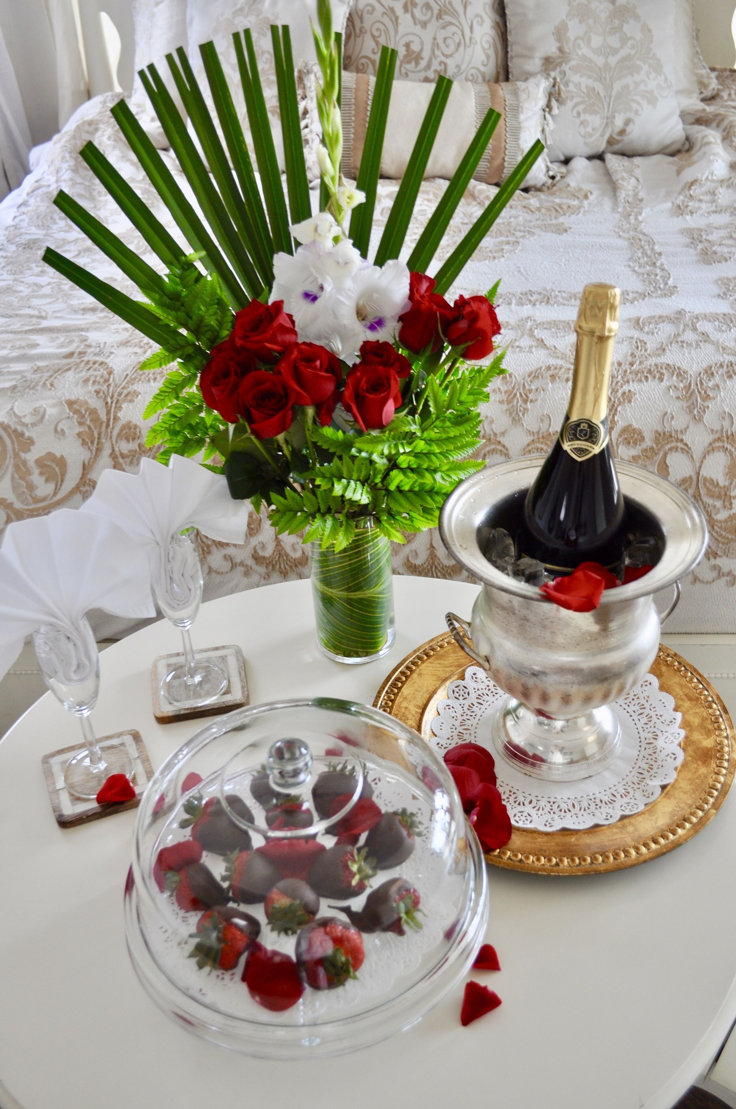 A table with a bouquet of red and white flowers, a glass of champagne, and a chocolate-covered strawberries dish.