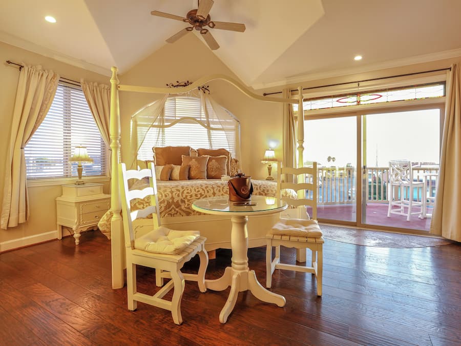 A bedroom with a four-poster bed, a round table, two chairs, a ceiling fan, and a balcony with outdoor furniture.