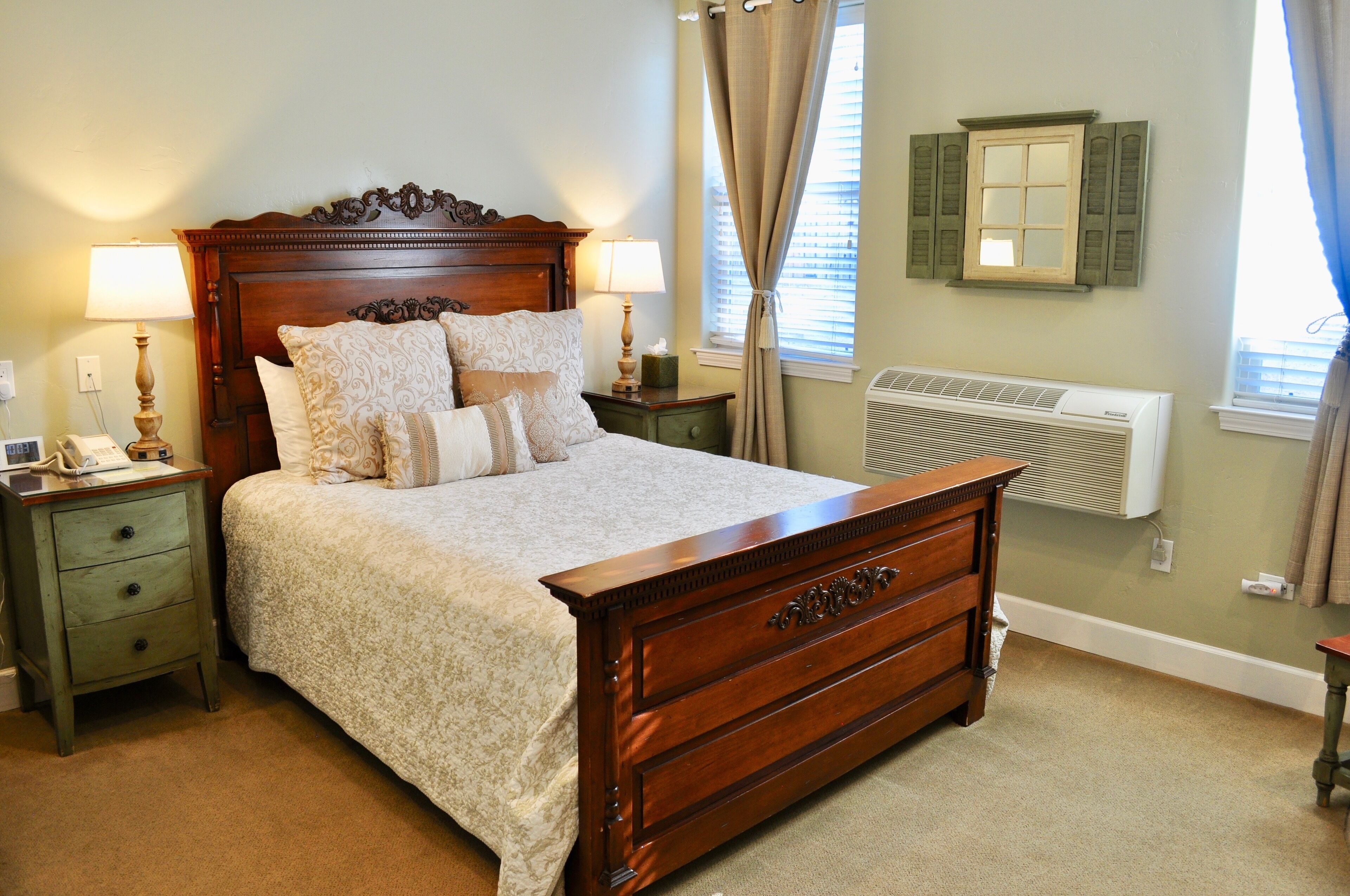A bedroom with a wooden bed, two bedside tables with lamps, a nightstand, a window with shutters, and a wall-mounted air conditioner.