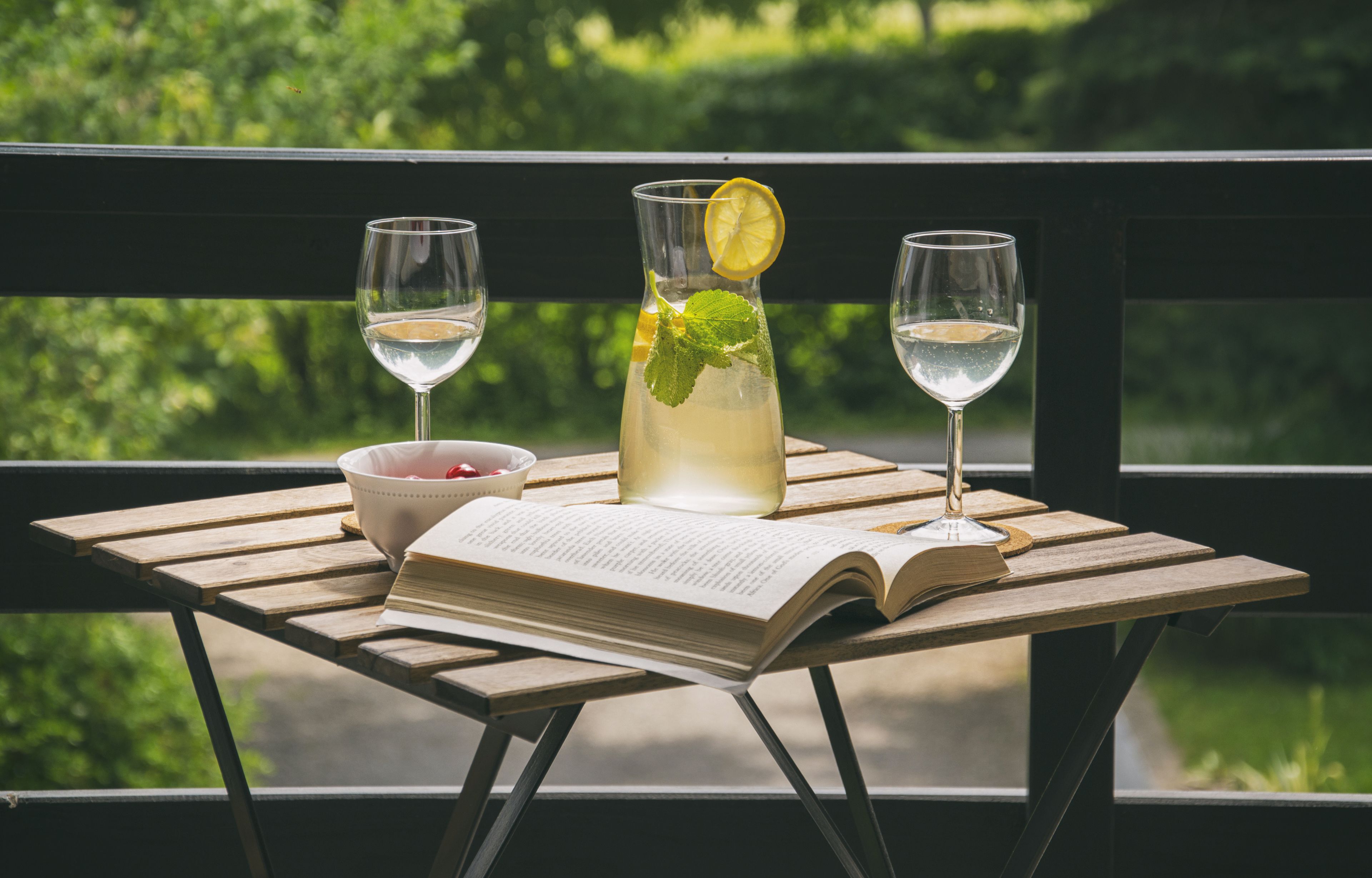 A wooden table with a book, a glass of white wine, a glass of a clear beverage with a lemon slice, and a bowl of fruit on a balcony with a view of greenery.