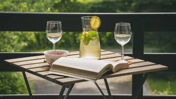 A wooden table with a book, a glass of white wine, a glass of a clear beverage with a lemon slice, and a bowl of fruit on a balcony with a view of greenery.