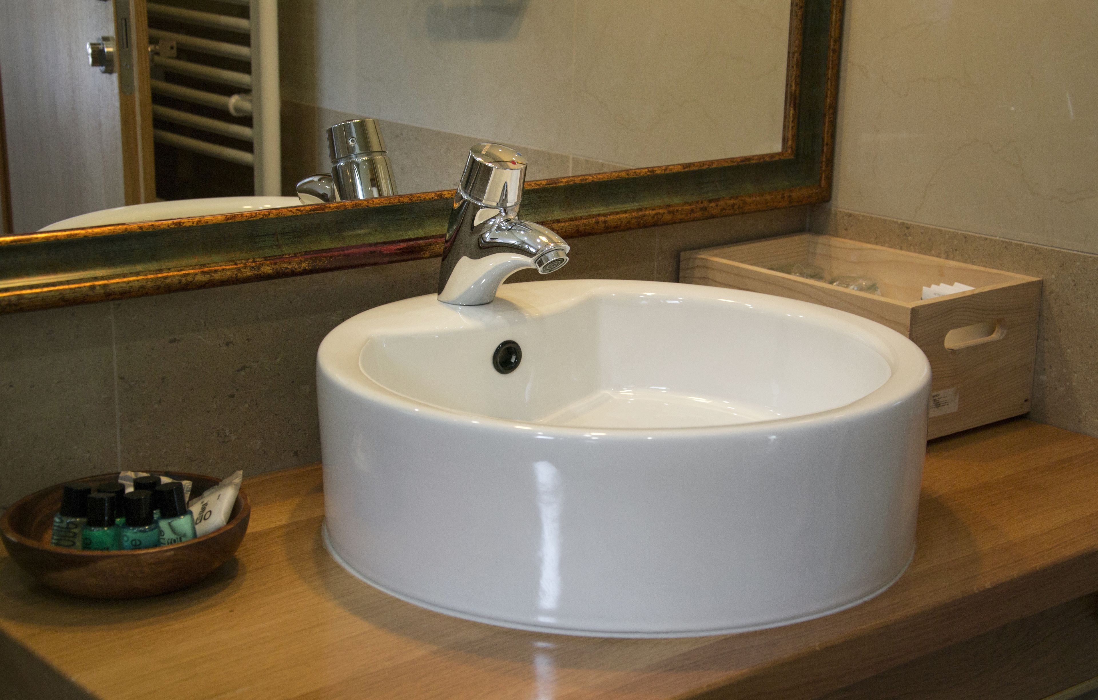 Un baño con un lavamanos blanco, un cuenco de madera con artículos de aseo y un espejo con marco decorativo.