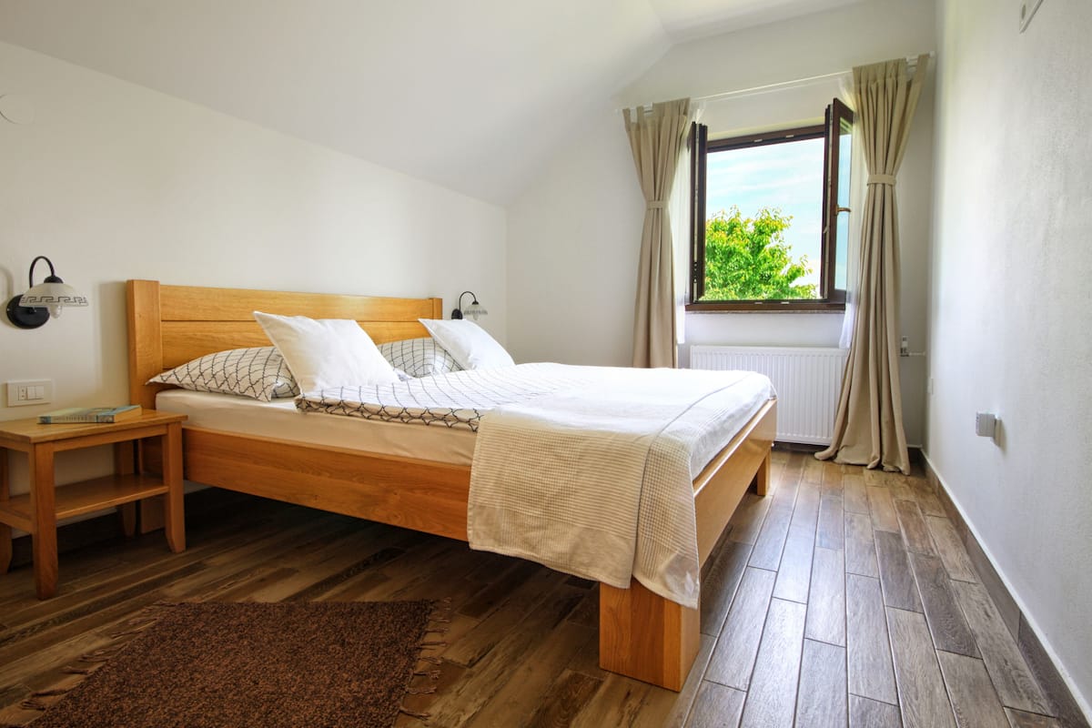 A bedroom with a wooden bed, a bedside table, a nightstand, a window with curtains, and a view of greenery outside.