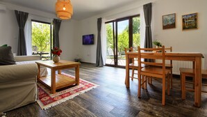 A spacious living area with a sofa, wooden coffee table, and dining table with chairs. There is a large window with curtains, a television, and artwork on the wall. - Lagom Apartments (Rakovica)