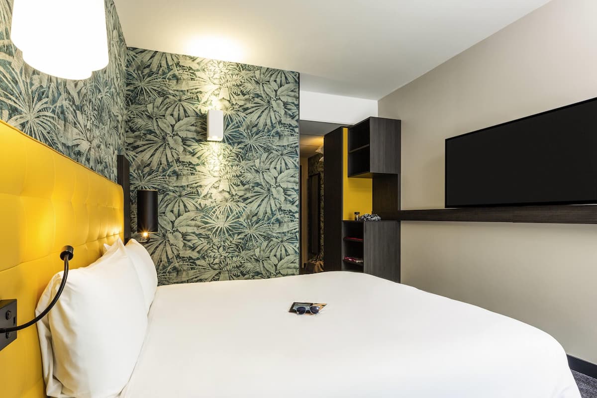 A hotel room with a bed, a mounted TV, a wall-mounted lamp, and a patterned wallpaper.