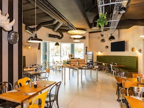 Um restaurante com várias mesas, cadeiras, um banco estofado verde e iluminação pendente, além de uma grande janela que oferece vista para o exterior.