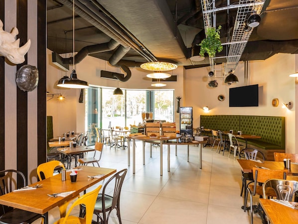 A restaurant with multiple dining tables, chairs, a green cushioned bench, pendant lighting, and a large window providing a view of the outdoors.