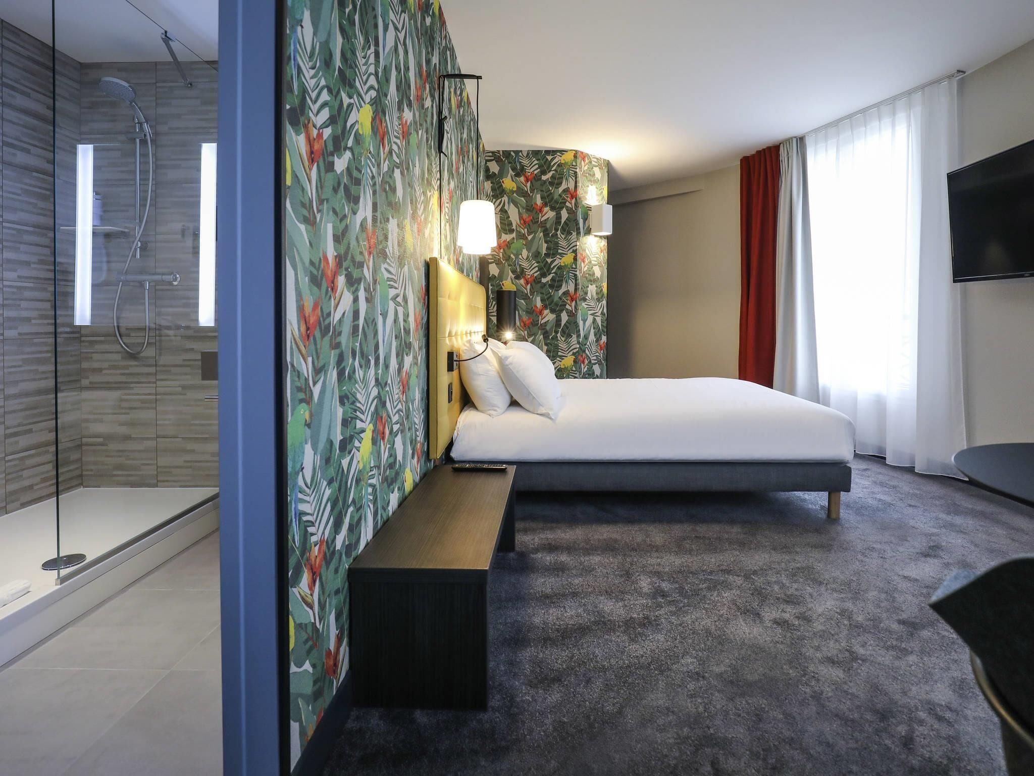 A modern hotel room with a shower, a bed, a bench, a TV, and a large window.