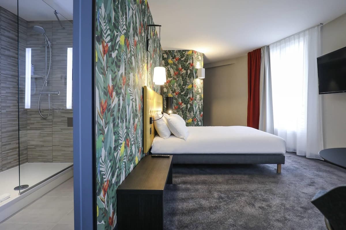 A modern hotel room with a shower, a bed, a bench, a TV, and a large window.