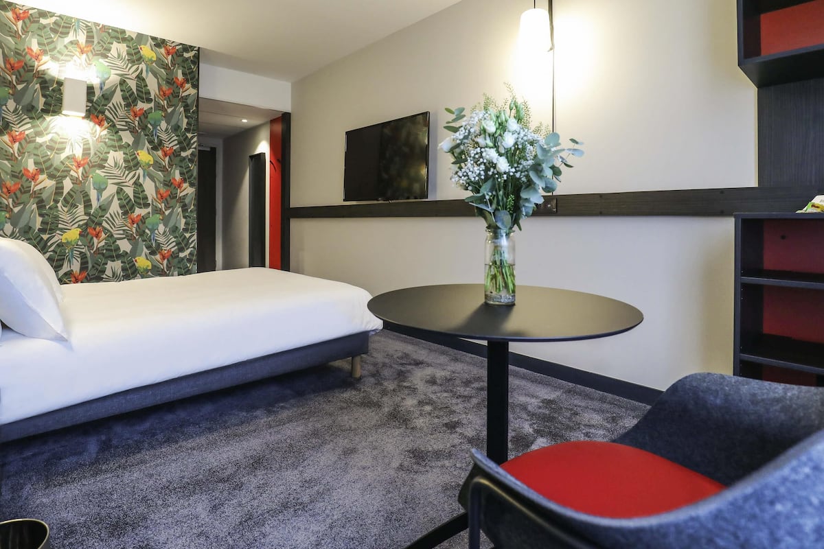 A hotel room with a bed, a round table with a vase of flowers, a chair with a red cushion, and a wall with a tropical leaf pattern.