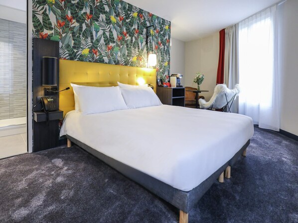 Habitación de hotel con una cama grande, cabecera con detalle amarillo, escritorio con lámpara, una silla y ventana con cortinas.